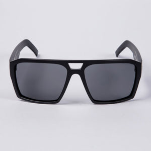The UNIT Vault Polarised Eyewear is showcased against a plain white background, featuring black sunglasses with a thick, angular frame and dark polycarbonate polarized lenses. The bridge of the sunglasses sports a distinctive triangular gap at the top for added style.