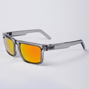 The UNIT Primer Polarised Eyewear showcases a pair of transparent gray sunglasses with reflective orange-red lenses. The thick temple arms feature the word "UNIT" in capital letters near the hinges. Boasting a lightweight frame and polarized lenses offering UV protection, these sunglasses rest stylishly on a white surface.