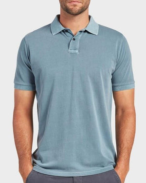 A man wearing an Academy Brand Academy Polo stands against a plain background. His light blue, short-sleeved cotton pique shirt features a buttoned collar and is untucked, with his hands partially in his pockets. The person's head is not fully visible in the image.