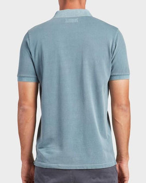 A person wearing a light blue short-sleeved Academy Brand Academy Polo stands with their back facing the camera. The comfortable polo has a collar, and the person is also wearing dark-colored pants. The background is plain and light-colored.