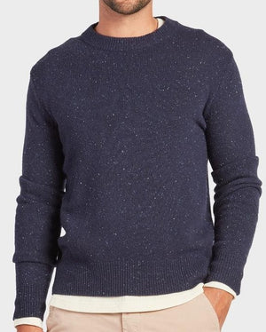 A man wears the Academy Brand Shoreditch Crew Knit, a navy speckled lambswool jumper with a crew neck, layered over a light shirt and paired with beige pants. He stands with one hand in his pocket, face partially out of frame.