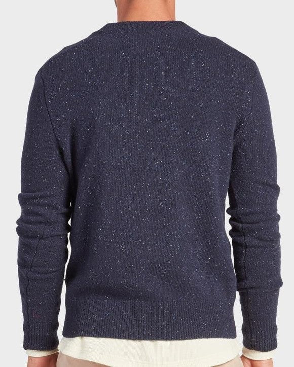 A person is shown from the back wearing the Academy Brand Shoreditch Crew Knit, a dark blue speckled lambswool jumper, over a white shirt and beige pants against a plain light background.
