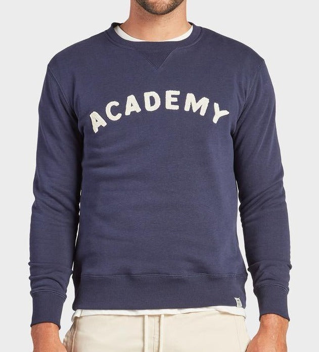 Someone wears the Academy Brand College Crew in navy, featuring bold white "ACADEMY" lettering across the chest, paired with light pants for a casual look against a plain light gray background.