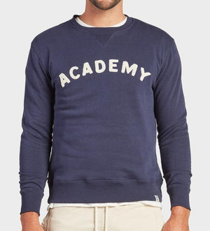 Someone wears the Academy Brand College Crew in navy, featuring bold white "ACADEMY" lettering across the chest, paired with light pants for a casual look against a plain light gray background.