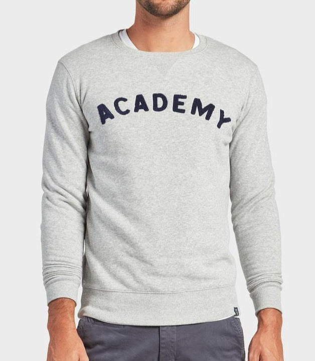 A man wears an Oxford Heather Academy Brand College Crew sweatshirt with "ACADEMY" in bold black letters on the chest, paired with gray pants. The background is plain and light-colored.