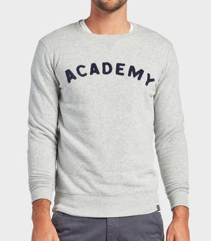 A man wears an Oxford Heather Academy Brand College Crew sweatshirt with "ACADEMY" in bold black letters on the chest, paired with gray pants. The background is plain and light-colored.