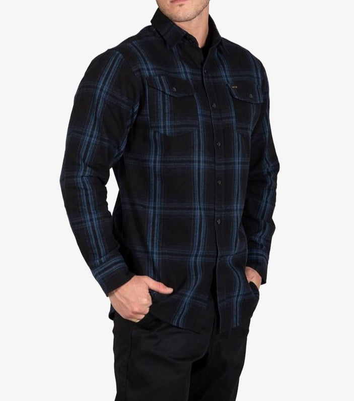 A person wearing a UNIT Newtown Flannel Shirt, characterized by its dark blue and black hues and featuring two buttoned chest pockets—one adorned with a small tag. The 100% cotton flannel shirt is paired with black pants, while the individual's hands are tucked into their pockets as they stand against a plain white background.