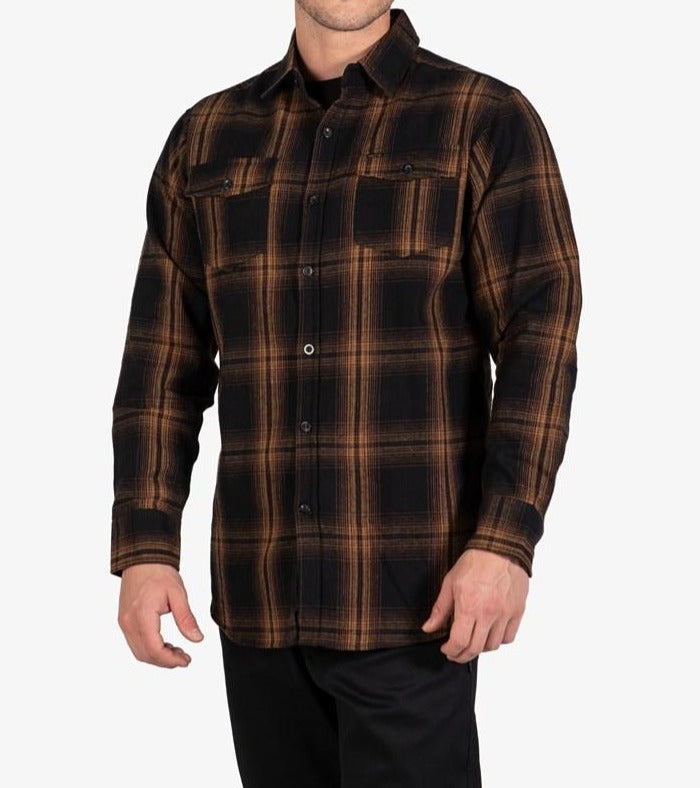 A person wearing the UNIT Newtown Flannel Shirt, made from 100% cotton in a black and brown plaid pattern, featuring two buttoned chest pockets. The shirt has long sleeves, and the person is standing with their hands relaxed at their sides. It is paired with black pants against a plain white background.