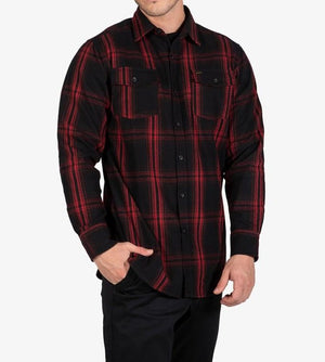 A person is wearing the UNIT Newtown Flannel Shirt, which features a black and red checkered pattern, is made from 100% cotton, and has two buttoned chest pockets along with long sleeves. The individual stands with one hand in their pocket, showcasing the shirt's design against a plain white background.