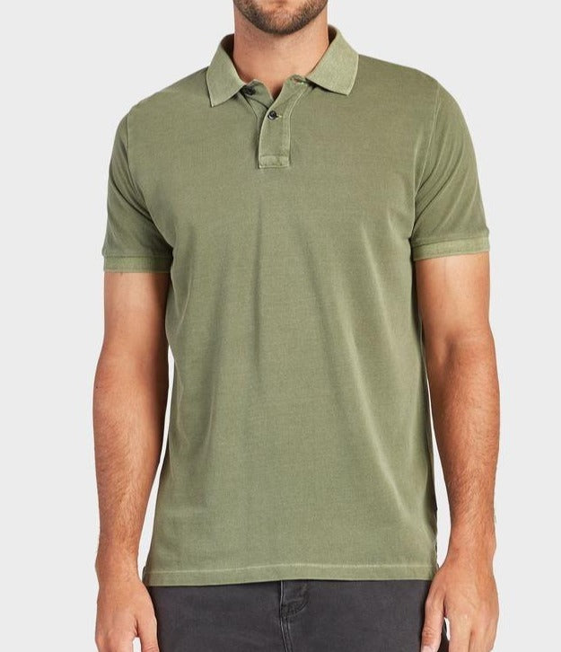 A man is wearing the Academy Brand Academy Polo, a short-sleeved, olive-green shirt featuring a collar and three buttons at the neck. Made of cotton pique for maximum comfort, he stands against a neutral background and pairs it with dark-colored pants.
