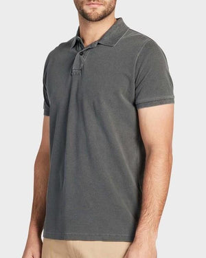 A person in an Academy Brand Academy Polo, a dark gray and comfortable cotton pique shirt with a collar and three-button placket, stands against a plain gray background. They are also wearing light khaki pants.