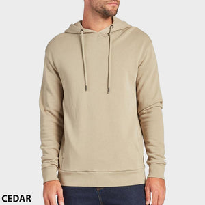 A man stands against a plain background wearing the Academy Brand Boston Hoodie in light beige with drawstrings and dark jeans. "CEDAR" is written in black text at the bottom left corner.