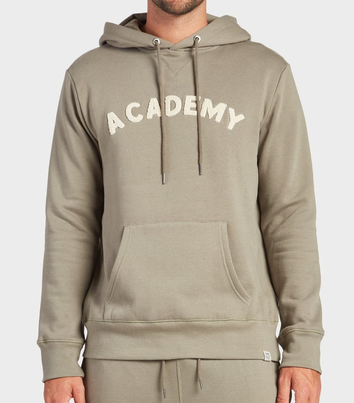 A person wearing an Academy Brand College Hoodie in moss, featuring "ACADEMY" in white textured letters across the chest, a front pocket, and drawstrings. The image shows the person from shoulders to hips.