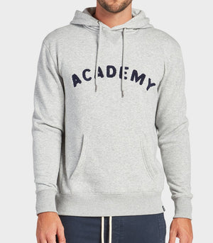 A man wears the Academy Brand College Hoodie in Oxford Heather, featuring "ACADEMY" in bold across the chest, a front pocket, and drawstrings. He pairs the casual sweatshirt with dark pants for a relaxed style.
