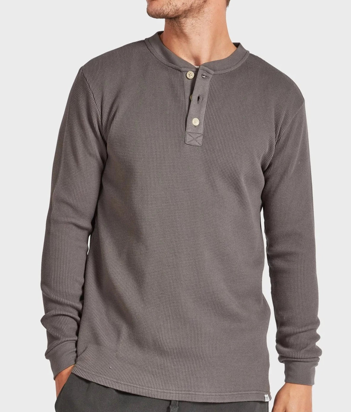 A man is wearing the stylish Academy Brand Sycamore Henley in dark gray, characterized by its waffle-knit texture and made from 100% cotton. This comfortable garment features a round neckline and a partial button-up placket with three buttons. He is also wearing dark pants and standing against a plain light gray background.