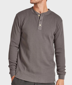 A man is wearing the stylish Academy Brand Sycamore Henley in dark gray, characterized by its waffle-knit texture and made from 100% cotton. This comfortable garment features a round neckline and a partial button-up placket with three buttons. He is also wearing dark pants and standing against a plain light gray background.