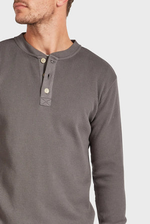 A person wearing the Academy Brand Sycamore Henley, a grey waffle-knit henley shirt made from 100% cotton, with three buttons partially undone. The person is turned slightly to the side, showcasing the shirt's texture and casual style against a plain white background.