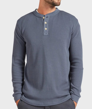 A man wearing the Academy Brand Sycamore Henley, a stylish, long-sleeve, blue waffle-knit shirt made from 100% cotton with three buttons. His face is not visible as he poses casually with his hands in his pockets against a plain white background.