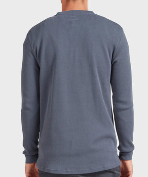 A person wearing the Academy Brand Sycamore Henley, a long-sleeve, 100% cotton shirt with a textured blue finish and subtle pattern, is shown from the back against a plain, light background.