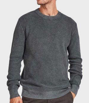 A man in the Academy Brand Newport Crew Knit—a textured dark gray 100% cotton, long-sleeve jumper—stands with one hand in his pocket against a plain light background, head slightly turned and face partially out of frame.