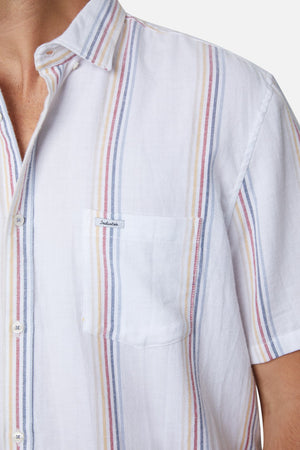Close-up of a person wearing the Industrie The Zante S/S Shirt, a comfortable, 100% cotton short-sleeved shirt featuring vertical stripes in blue, red, and yellow. This casual shirt includes a front pocket on the left side with a small white label displaying text. The background is plain and the person's head is partially visible.