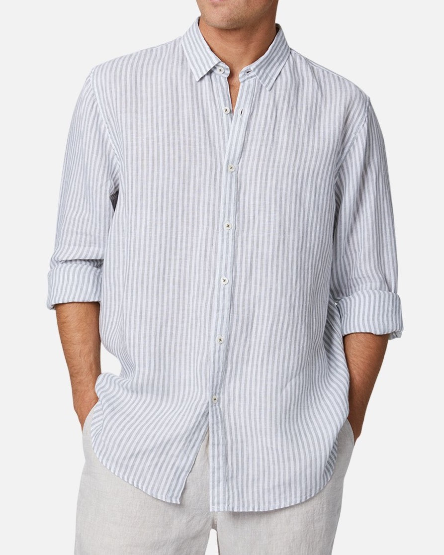 A man wearing the Industrie The Southwest Linen L/S Shirt, featuring light blue and white vertical stripes with rolled-up sleeves, paired with light-colored pants and standing with his hands in his pockets for a relaxed look.