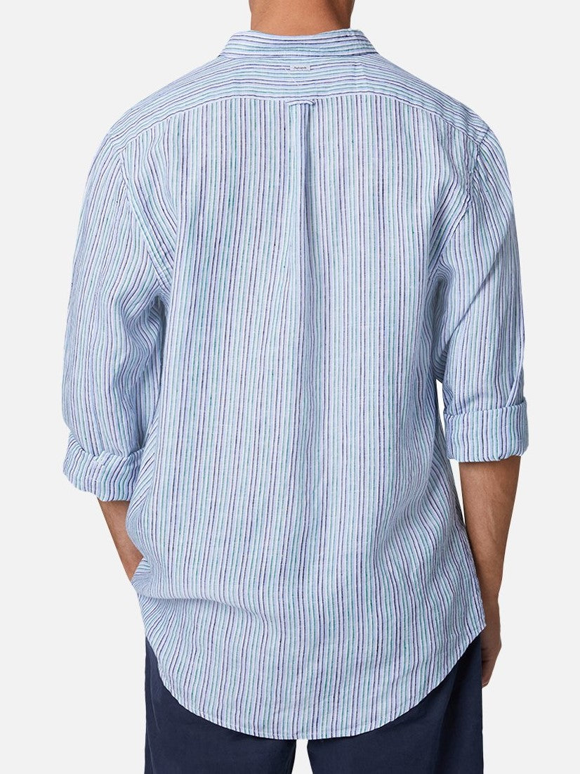 A person wearing the Industrie The Grenadines Linen Shirt in light blue, a long-sleeve, vertically striped linen shirt with rolled-up sleeves, viewed from the back. The relaxed fit shirt is untucked and paired with dark shorts.