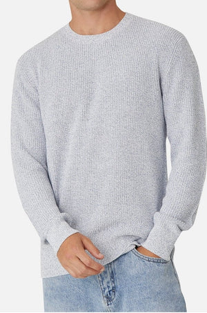 A man is wearing a light gray Industrie The Kingston Knit, paired with light blue jeans. He stands against a plain white background, casually placing one hand in his pocket.