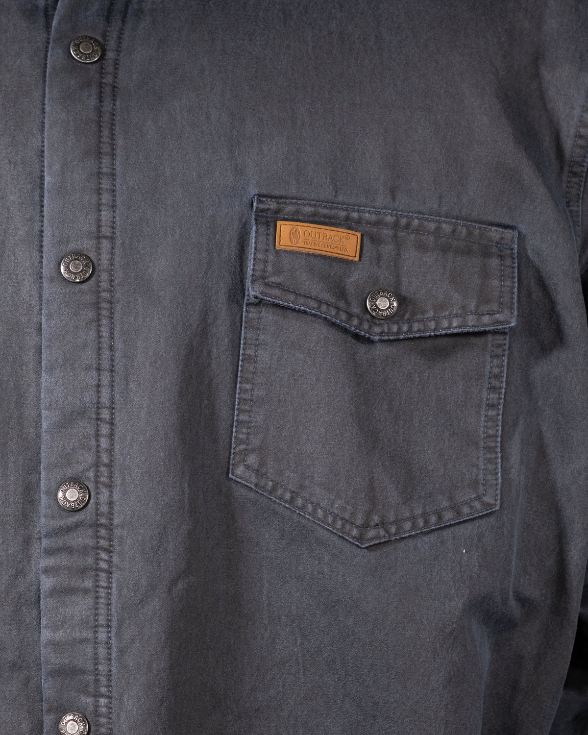 Close-up of the Outback Loxton Jacket in dark gray, highlighting a chest pocket with a small brown leather patch featuring indistinct text. This shirt-jacket is made from textured fabric with fleece lining for extra comfort.