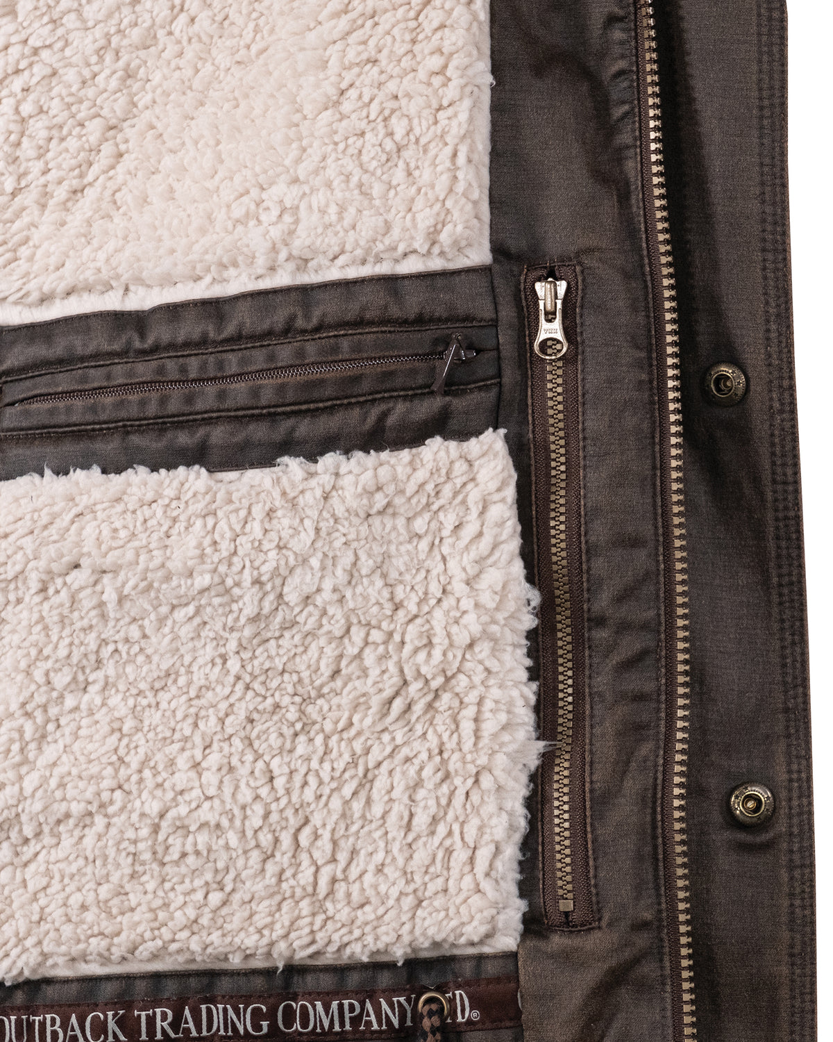 Close-up of the interior of a brown Outback Nolan Jacket, highlighting a front zipper on the right side and an interior pocket with a zipper on the left. The lining of this comfortable jacket is made of white faux sherpa material. The brand "OUTBACK TRADING COMPANY" is partially visible at the bottom.