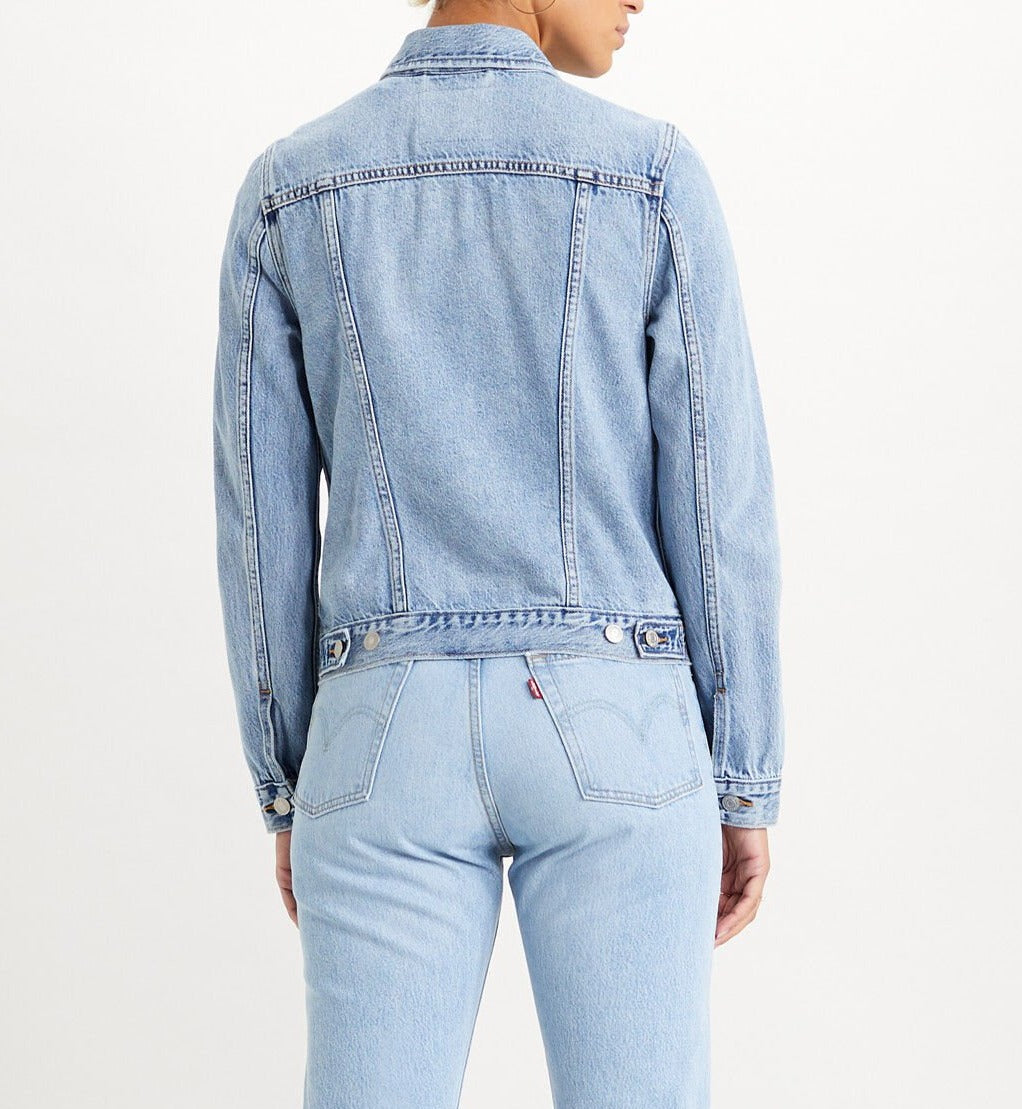 A person wearing a Levis Women's Original Trucker Jacket in light blue denim paired with matching jeans stands against a solid off-white backdrop. The jacket, adorned with stitching details and buttoned cuffs, complements the classic-fit jeans for an iconic wardrobe staple look.