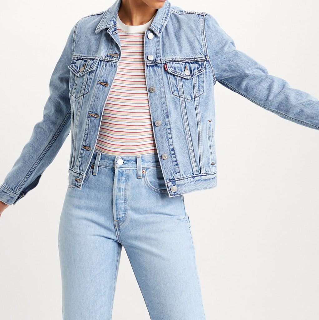 Styled in a Levi's Women's Original Trucker Jacket in light blue denim over a colorful striped t-shirt, they pair it with matching jeans. With arms outstretched against a plain white background, this wardrobe staple is showcased effortlessly.