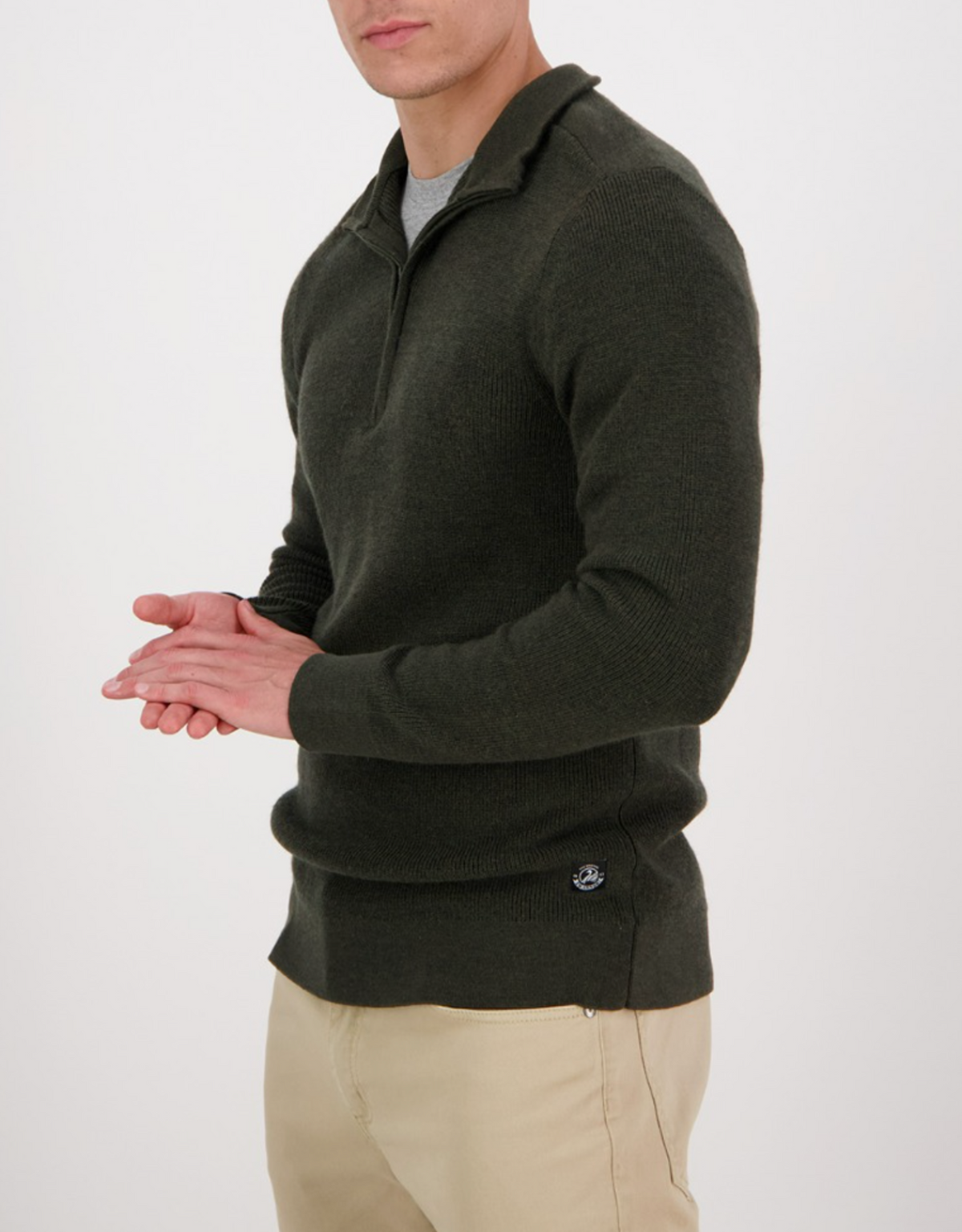 A person in a dark green Swanndri Magnet Bay v2 Merino Jumper with a half zip collar and beige pants stands against a plain white background, their hands clasped together.