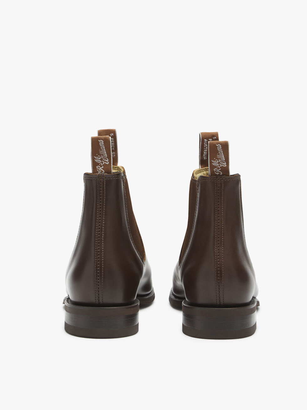 The RM Williams Comfort Craftsman Boot is depicted from the back, showcasing handcrafted brown leather Chelsea boots with pull tabs adorned with white text and dark wooden heels. These premium yearling leather boots feature visible stitching along the back seam and include a comfort innersole for all-day wear, set against a plain white background.
