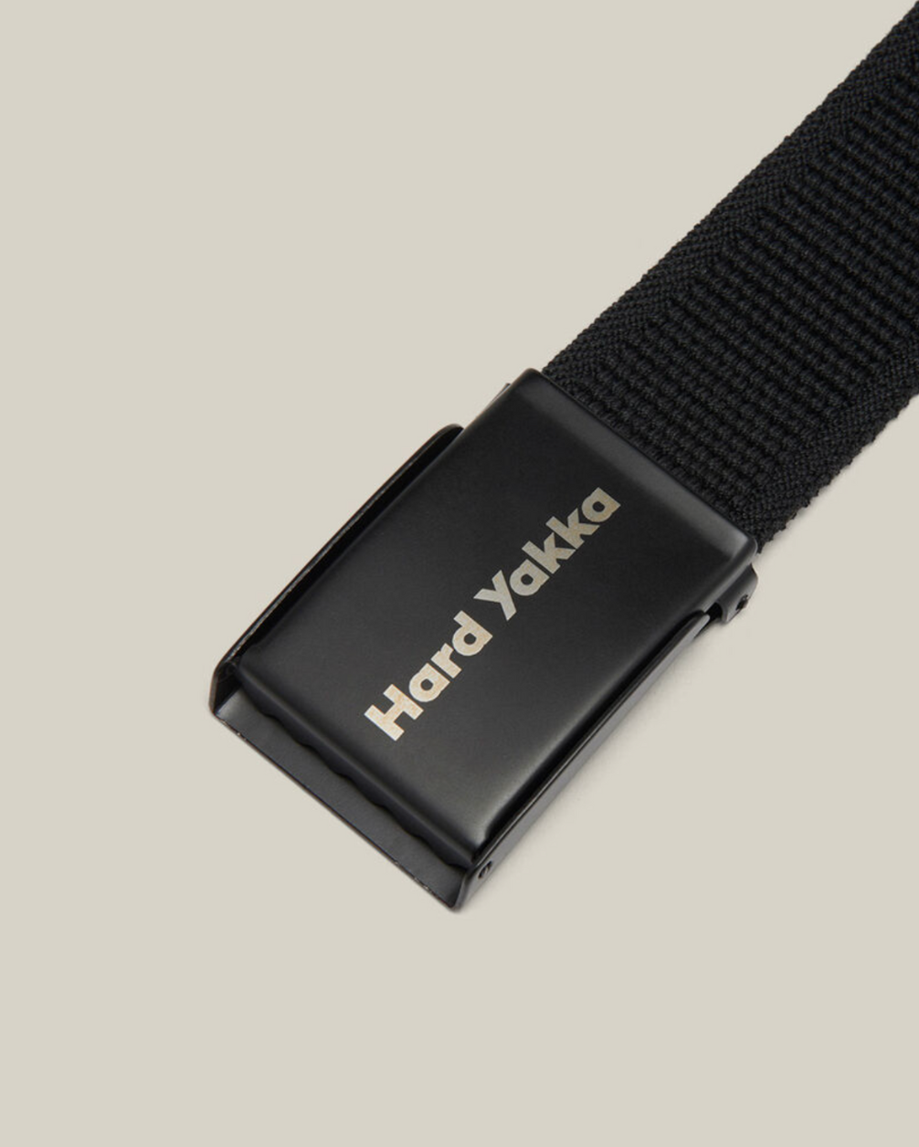 A close-up image of the Hard Yakka Stretch Webbing Belt in black, showcasing its sleek black metal buckle. The buckle displays the brand name "Hard Yakka" in subtle grey lettering and incorporates a hidden bottle opener. The belt is slightly angled against a simple beige background.