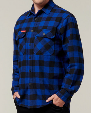 A person is wearing a Hard Yakka Check Flannelette Shirt with a blue and black checkered pattern. The durable, button-up cotton twill shirt features two front pockets with buttons, and the person has their hands in their pants pockets. The background is plain and light-colored.