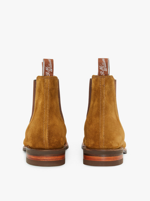 View of the RM Williams Comfort Suede Craftsman Boots from the back, showcasing pull tabs with white script lettering on a pair of handcrafted Australia brown suede Chelsea boots. These unworn boots feature ankle-height shafts, flat brown and tan stacked heels, and comfort rubber soles, all set against a plain white background.