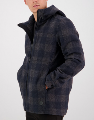 A person in a Swanndri Hudson Hoody, featuring a blue and gray plaid pattern, stands with one hand in their pocket. Made from New Zealand wool, the zipped jacket exudes a casual, warm vibe against a plain white background.