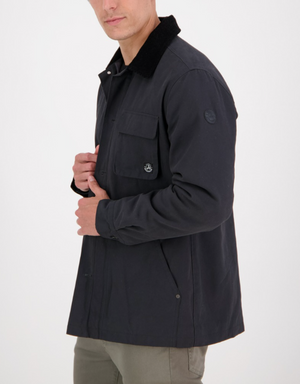A man models the Swanndri Barrytown v2 Canvas Work Jacket, a dark canvas jacket with a black collar, buttoned front, and two chest pockets. Featuring logos on the chest and sleeve, he stands against a white background, gently adjusting his jacket.