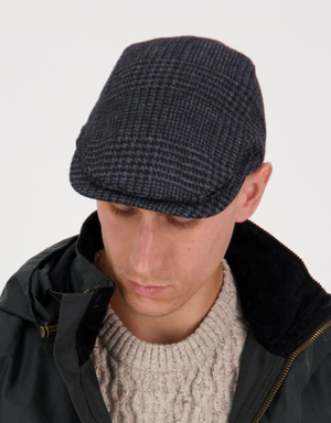 Someone wearing the dark Swanndri Bristol Flat Cap and a dark jacket over a cream sweater exudes classic style, looking down against a plain white background.