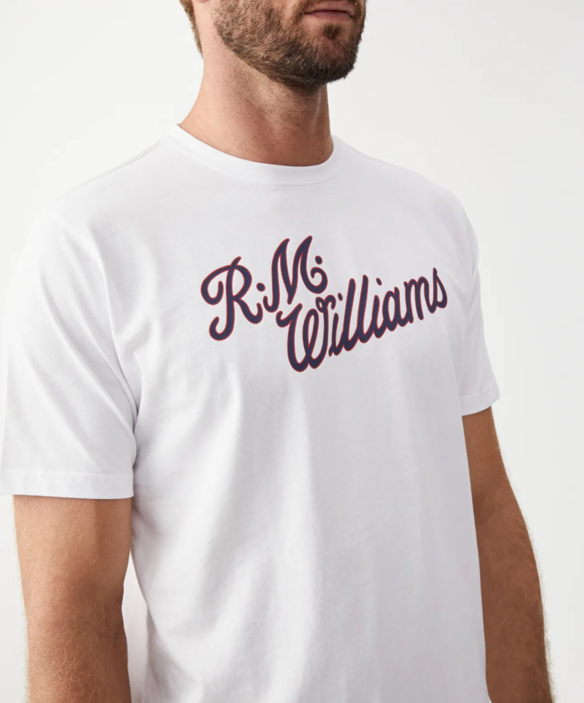 A man wearing a white RM Williams Script T-Shirt with "R.M. Williams" scripted in dark red across the chest. The image showcases the torso and front collar of this cotton shirt, with part of the man's face and beard visible against a plain white background.