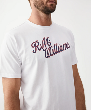 A man wearing a white RM Williams Script T-Shirt with "R.M. Williams" scripted in dark red across the chest. The image showcases the torso and front collar of this cotton shirt, with part of the man's face and beard visible against a plain white background.