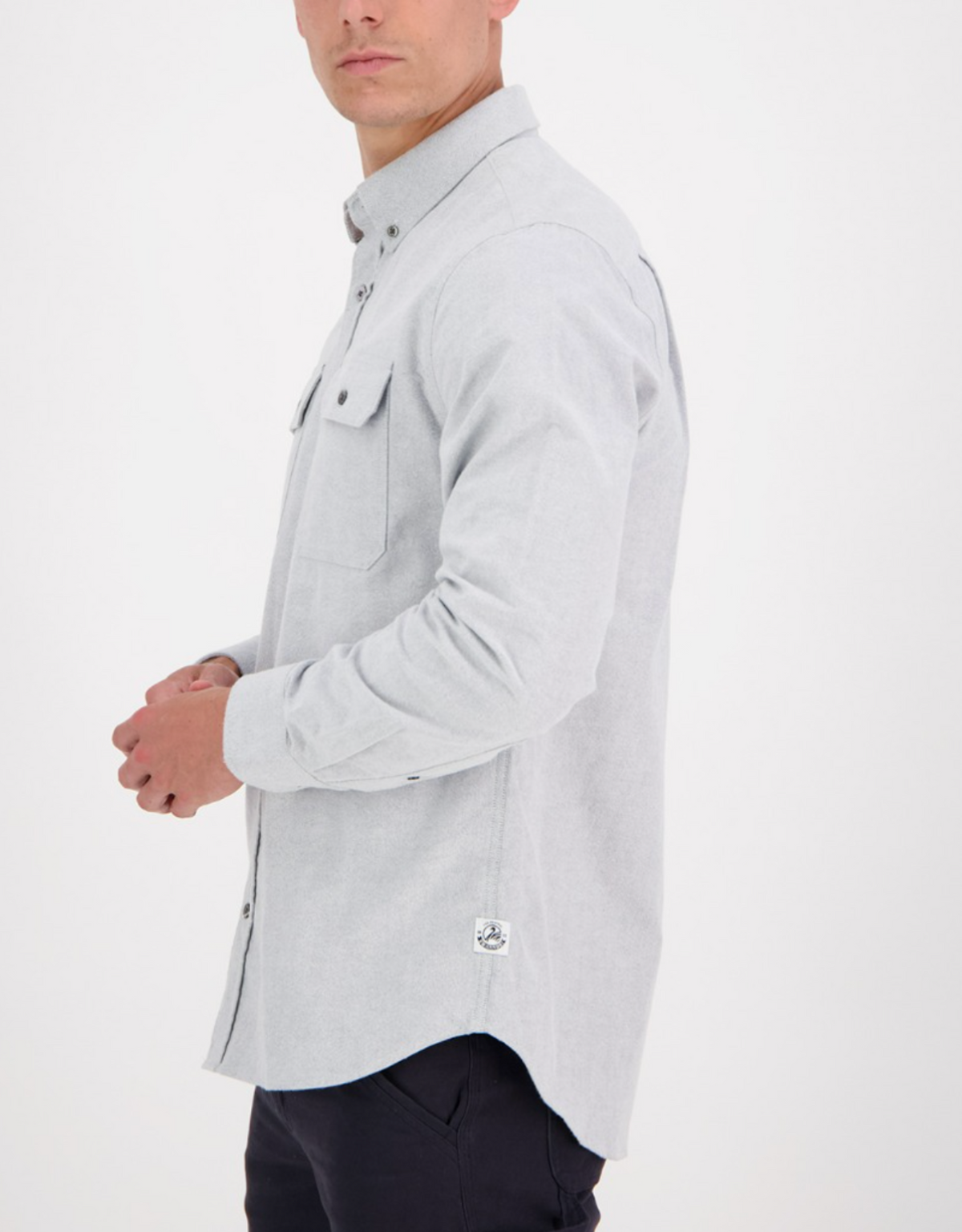 A person showcases the Swanndri Marwood Shirt, a stylish light gray long-sleeve button-up made from 100% cotton. It features two chest pockets and offers a classic fit. They stand sideways facing right, paired with dark pants against a white backdrop.