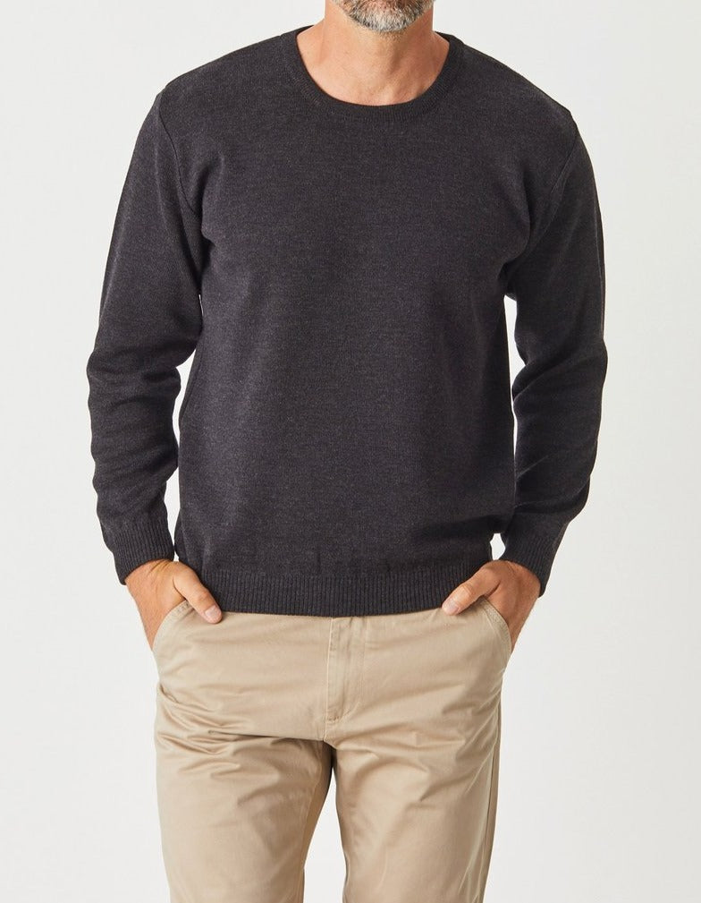 A man wears the Aklanda Crew Pullover Australian Made in dark gray with beige pants, standing hands-in-pockets against a plain white background. His head is partly visible, showing short hair and a beard.