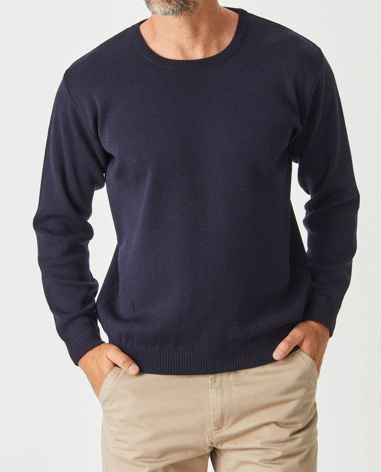 A man wears the Aklanda Crew Pullover Australian Made, a navy blue Merino Wool jumper, with beige pants. He stands with hands in pockets against a plain white background, his face partially out of frame.