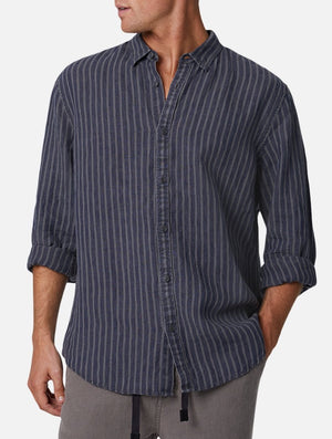 A man is wearing the Industrie The Chartres L/S Shirt, a dark blue, long-sleeved, casual shirt with vertical stripes and rolled-up sleeves. The 100% linen, relaxed fit shirt is paired with light gray pants. He stands with his hands in his pockets, looking to the side against a plain white background.
