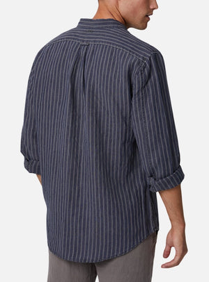 A person is standing with their back to the camera, wearing the Industrie The Chartres L/S Shirt, a long-sleeved, dark blue shirt with vertical stripes. The sleeves are rolled up and the shirt is untucked, displaying its relaxed fit. Made from 100% linen, this casual shirt is paired with grey pants against a plain white background.
