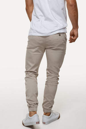 A person, back to the camera, wears a white T-shirt and light beige Industrie The Drifter Chino Pants with an elastic waist. White sneakers complete the outfit, offering a casual yet stylish vibe.