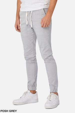 A person wearing Industrie The Drifter Chino Pant, a light gray with an elastic waist, a plain white T-shirt, and white sneakers. Their hands are relaxed by their side against a plain white background. "POSH GREY" is elegantly displayed in the bottom left corner.