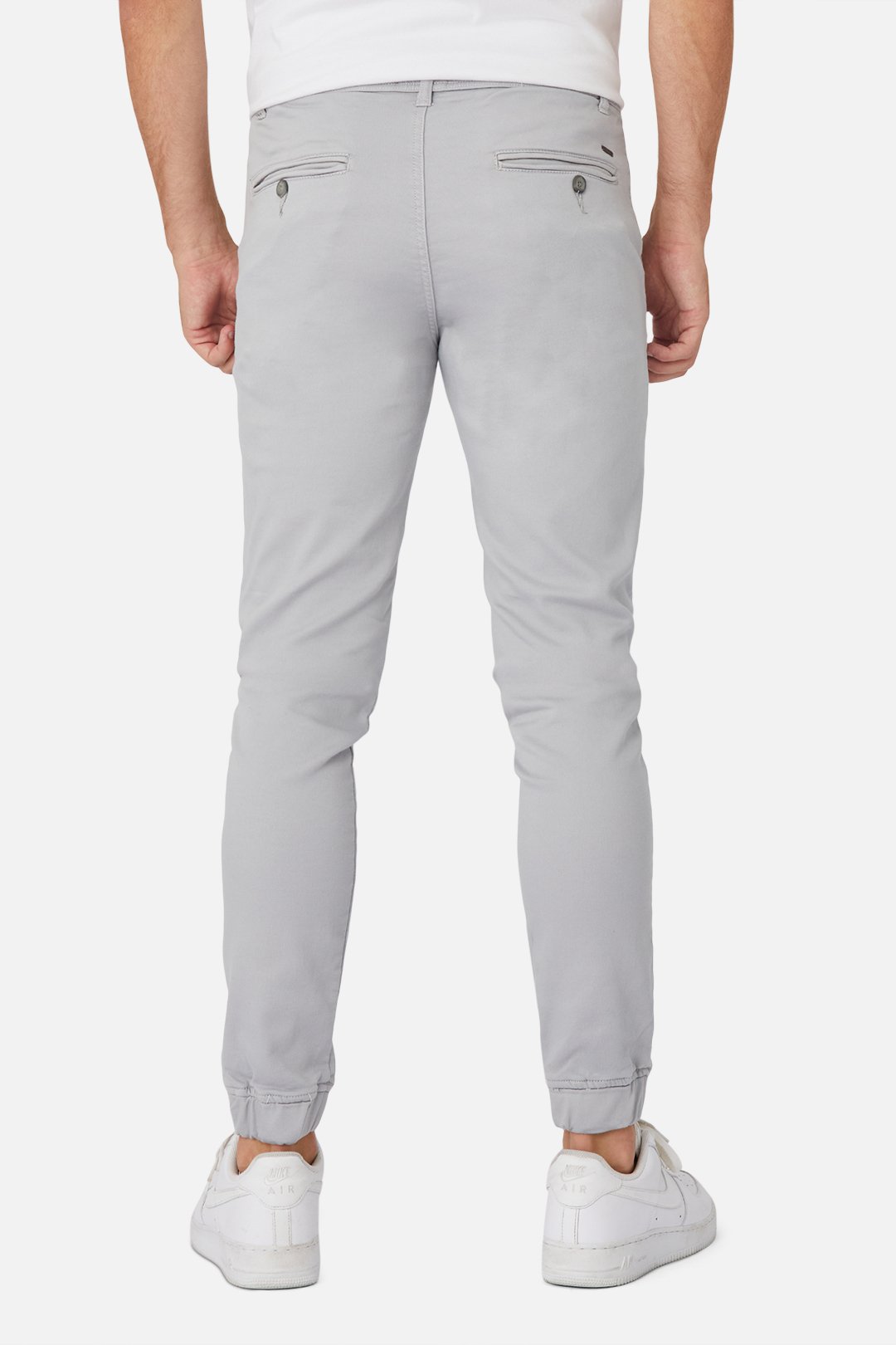 Someone is seen from the back wearing Industrie The Drifter Chino Pant in light gray, featuring a slim-fit jogger style with an elastic waist and buttoned back pockets, paired with white sneakers, set against a plain white background.